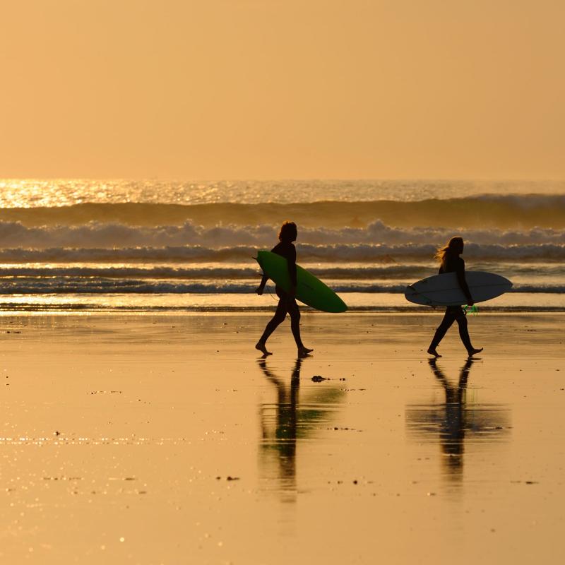 Two surfers carrying boards along Woolacombe beach with sea and sunset in background